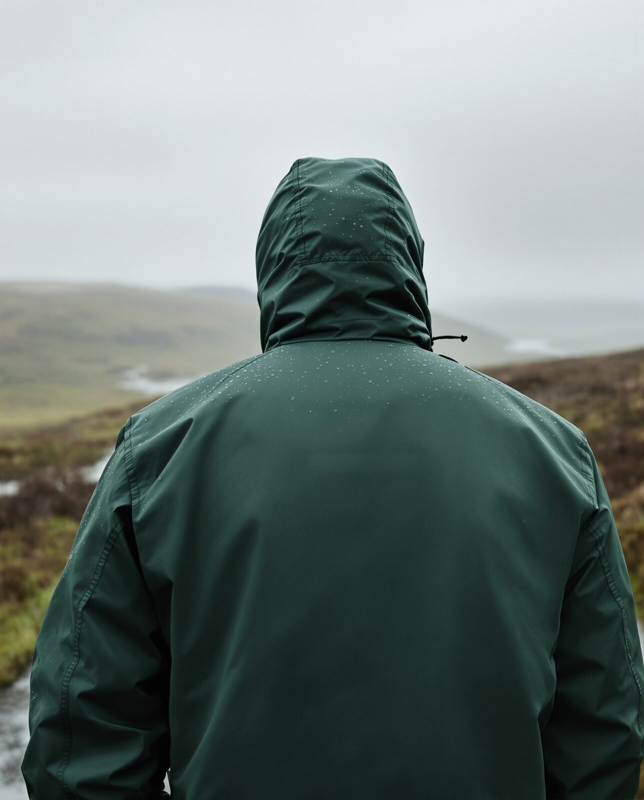 A walker in a green waterproof stands with the hood up on a wet moorland track.